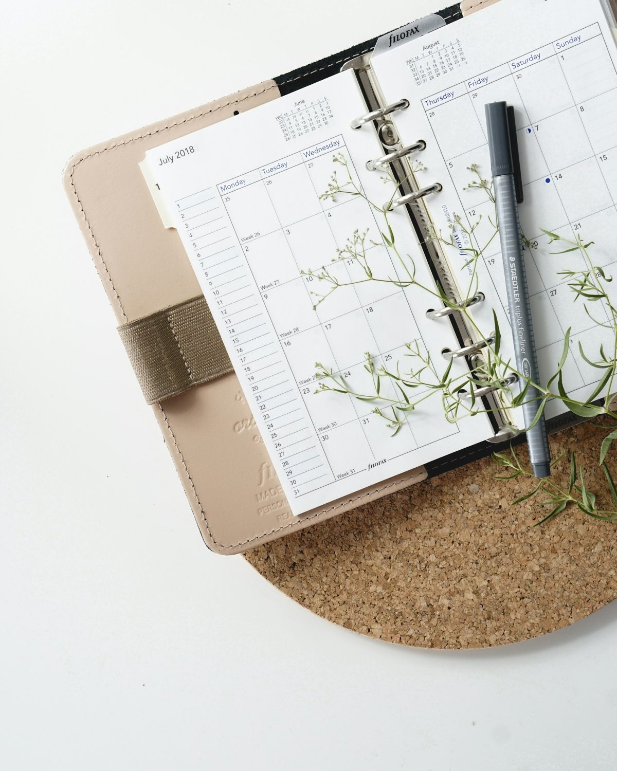 Open planner showing a calendar for July 2018 on a cork surface, with a black pen and sprigs of small white flowers.