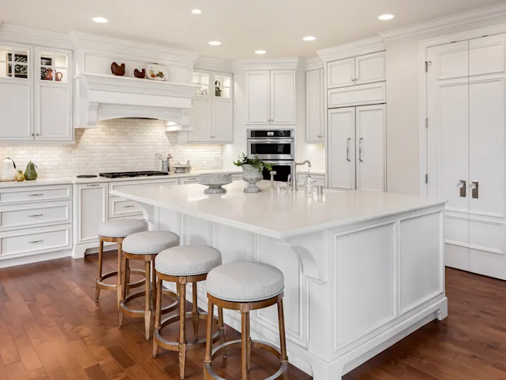 Spacious kitchen with white cabinets, an island with four stools, stainless steel appliances, and wooden flooring.