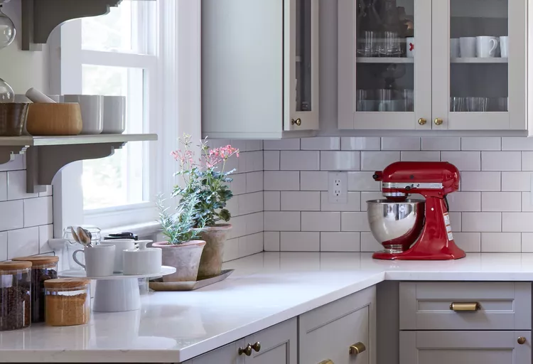 A kitchen corner with white cabinets, a subway tile backsplash, a red stand mixer, and plants on the countertop by a window.