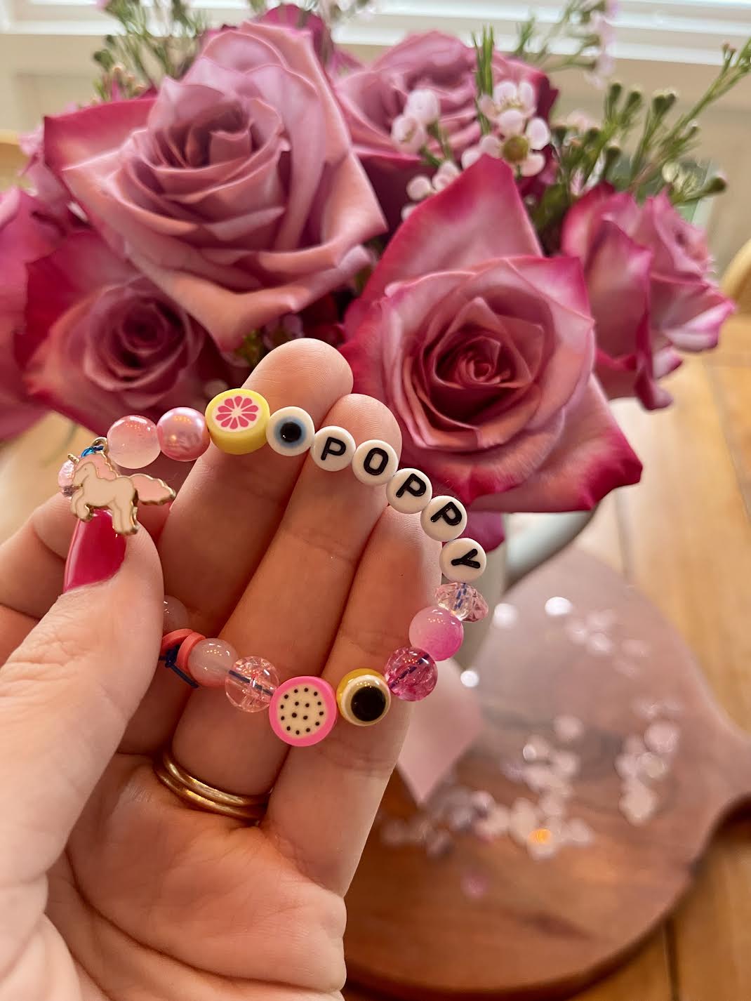 A hand holds a colorful beaded bracelet spelling "Poppy" in front of a bouquet of pink roses.