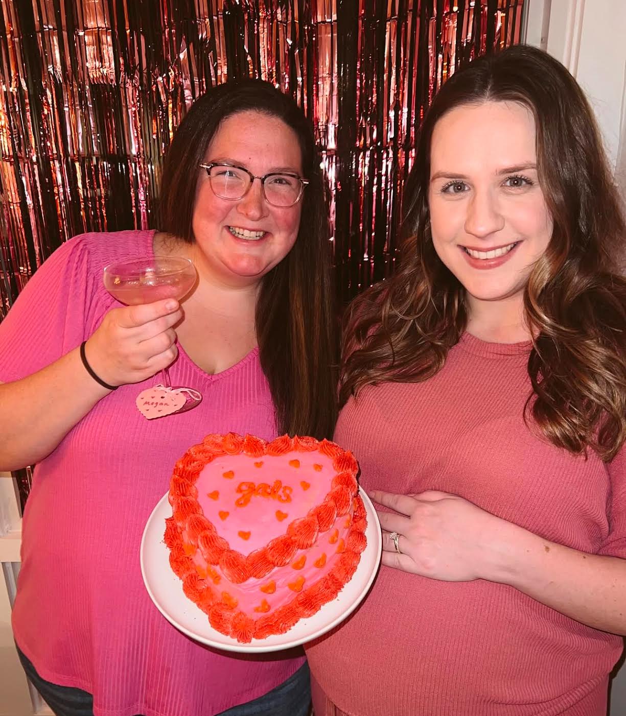 Two women smiling; one holds a heart-shaped cake with "gal's" on it, the other a drink. They stand in front of a metallic red backdrop.