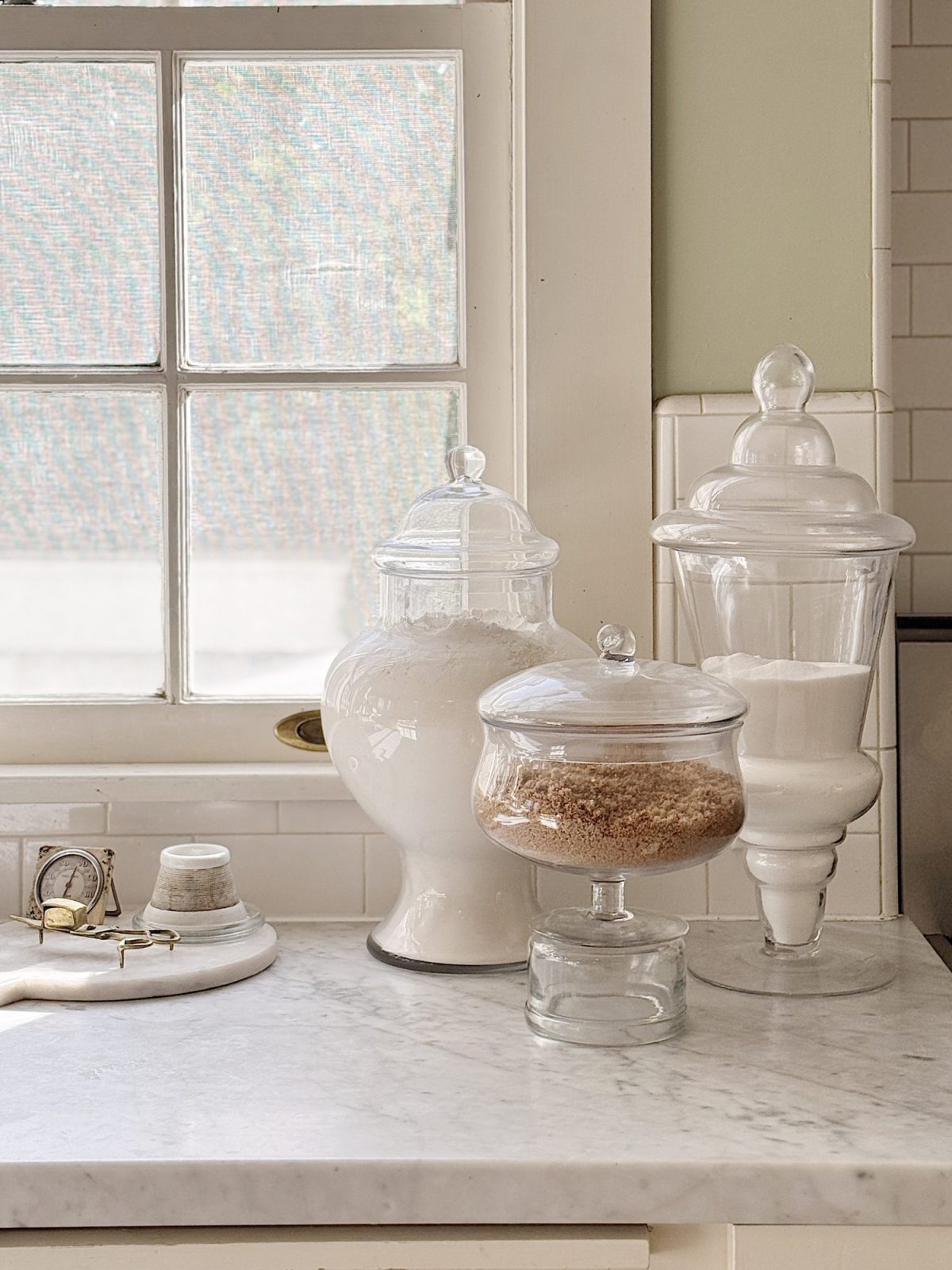 Three glass jars with lids containing flour and sugar are arranged on a marble countertop near a window, with a small marble tray holding a candle and match holder.