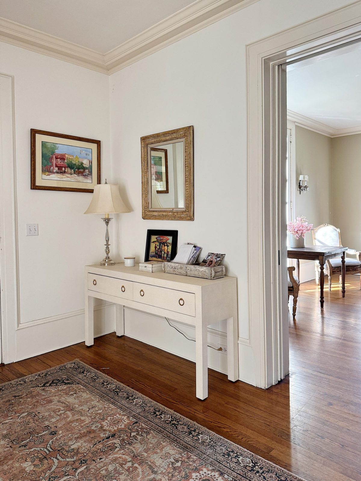 A hallway with a beige console table, a lamp, and framed pictures. Paintings hang above the table, and a rug covers the wooden floor. An open door leads to a light-filled room.