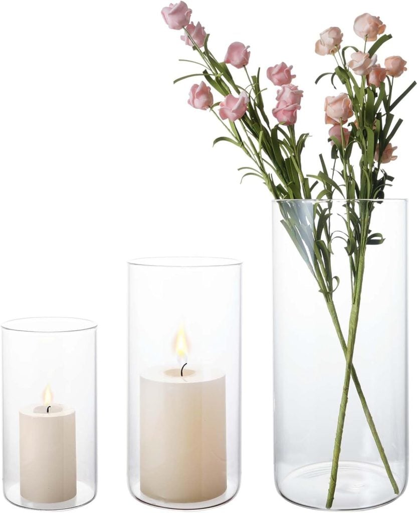Three clear glass cylindrical vases: the smallest holds a candle, the medium a larger lit candle, and the tallest—with flower frogs at the base—contains pink artificial flowers.