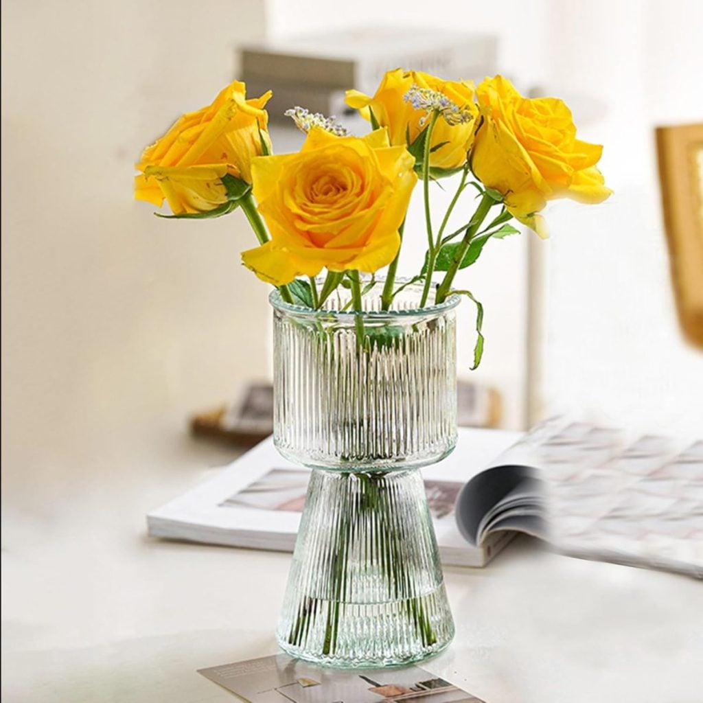 A clear ribbed glass vase holds five best faux flowers&mdash;yellow roses with green stems&mdash;placed on a white surface beside an open magazine.