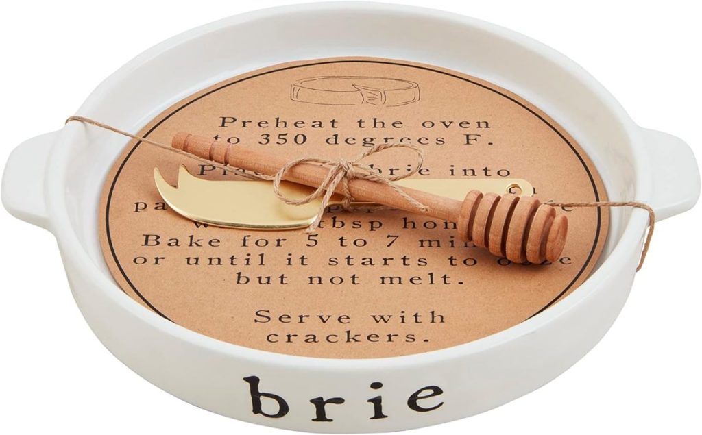 A white ceramic dish labeled &ldquo;brie&rdquo; holds a round of brie baking instructions, a gold cheese knife, and a honey dipper tied with string&mdash;perfect for preparing baked brie with jam.