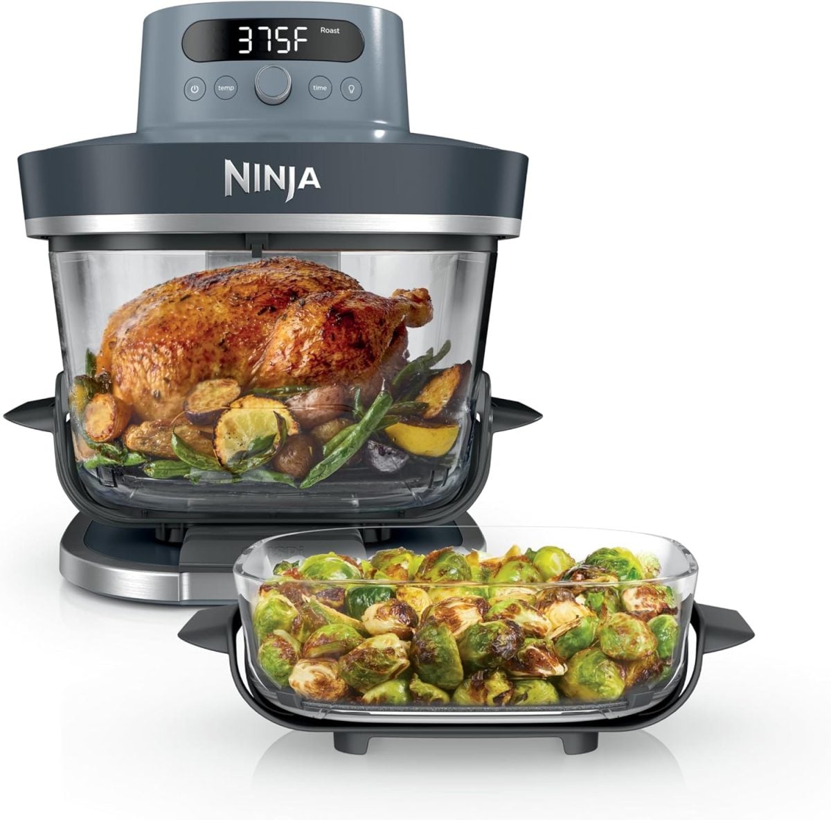 Ninja countertop oven cooking a whole baked parmesan crusted chicken with vegetables; a separate glass dish filled with roasted Brussels sprouts is in the foreground.