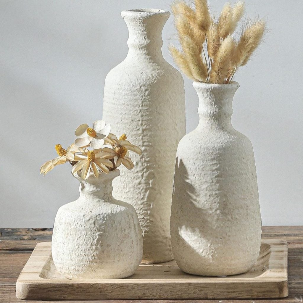 Three textured white ceramic vases on a wooden tray, with best faux flowers and grasses arranged in two of the vases, set on a wooden surface.