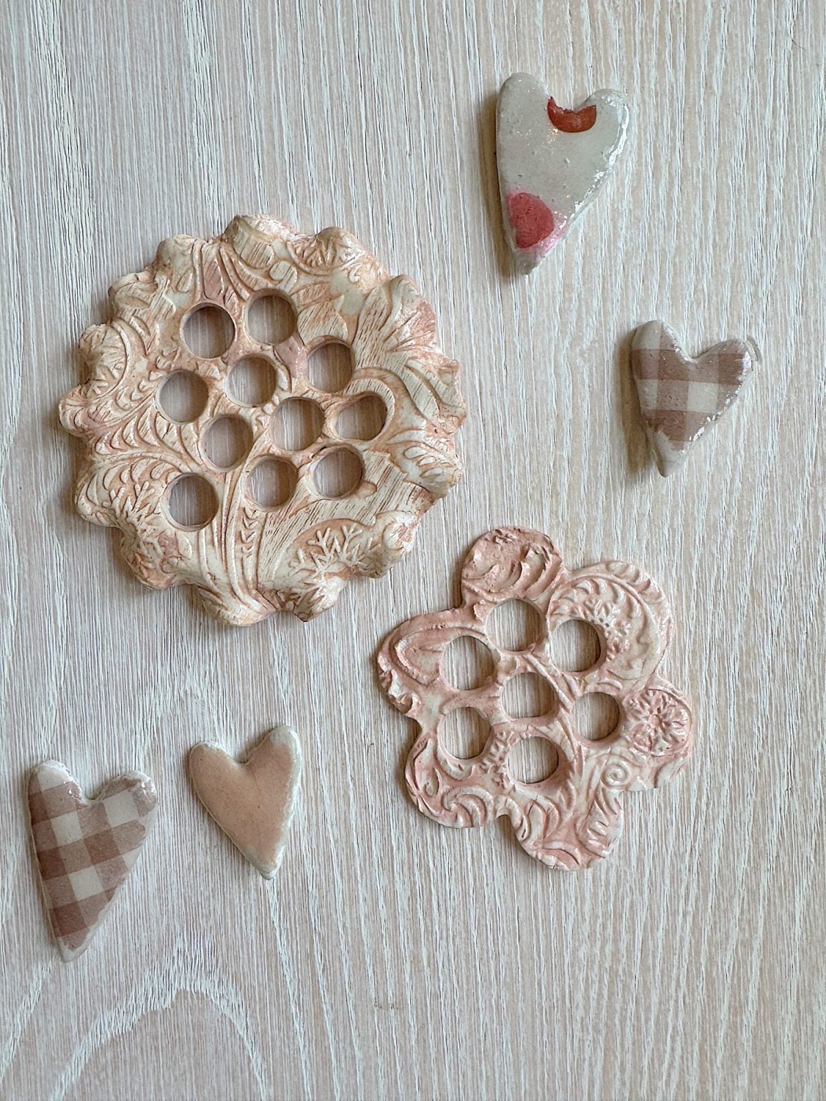 Ceramic buttons with floral cutouts and heart-shaped embellishments in various patterns and shades on a light wooden surface.