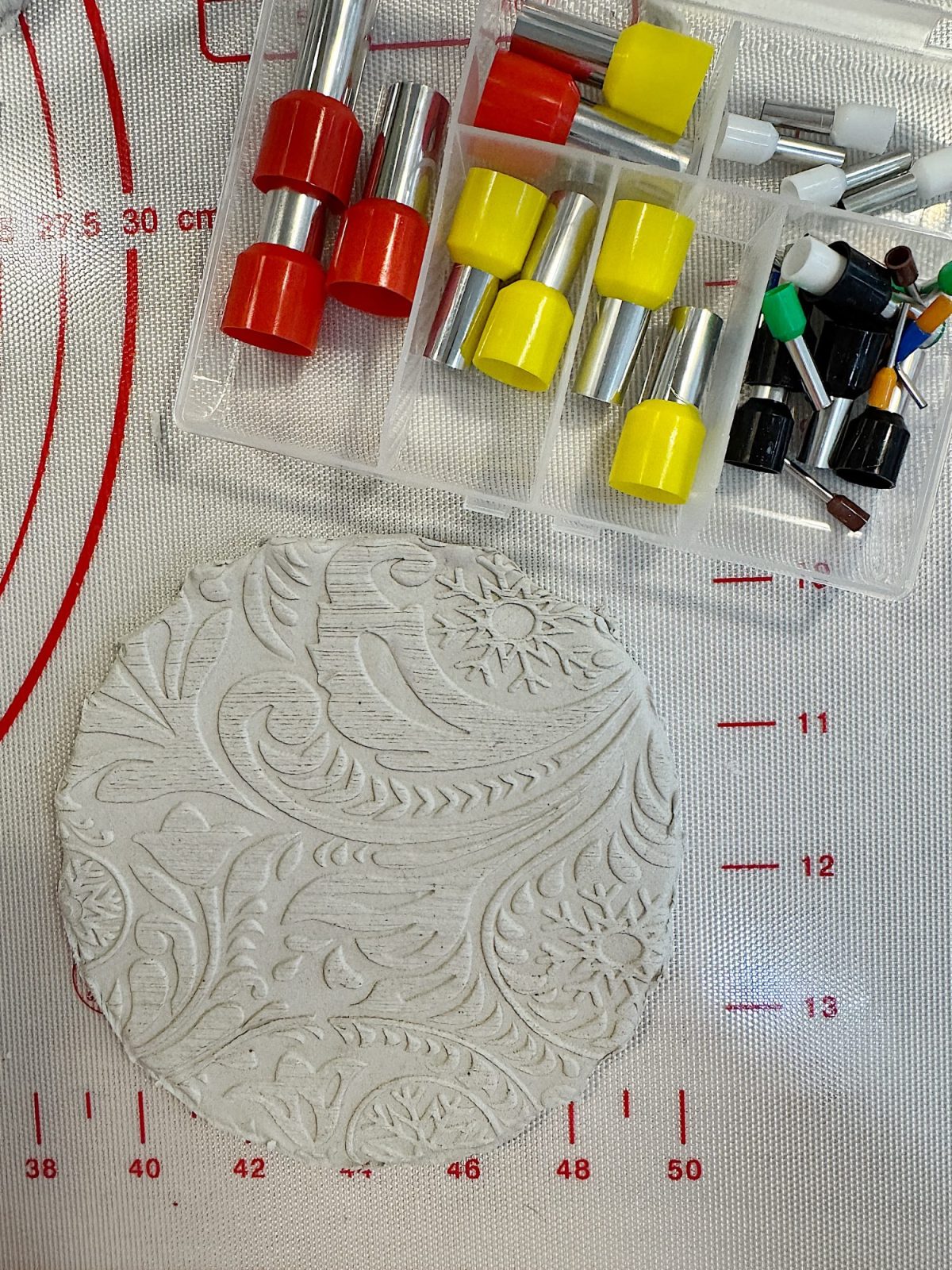 Embossed clay circle on a measuring mat with assorted metal and plastic clay tools beside it.