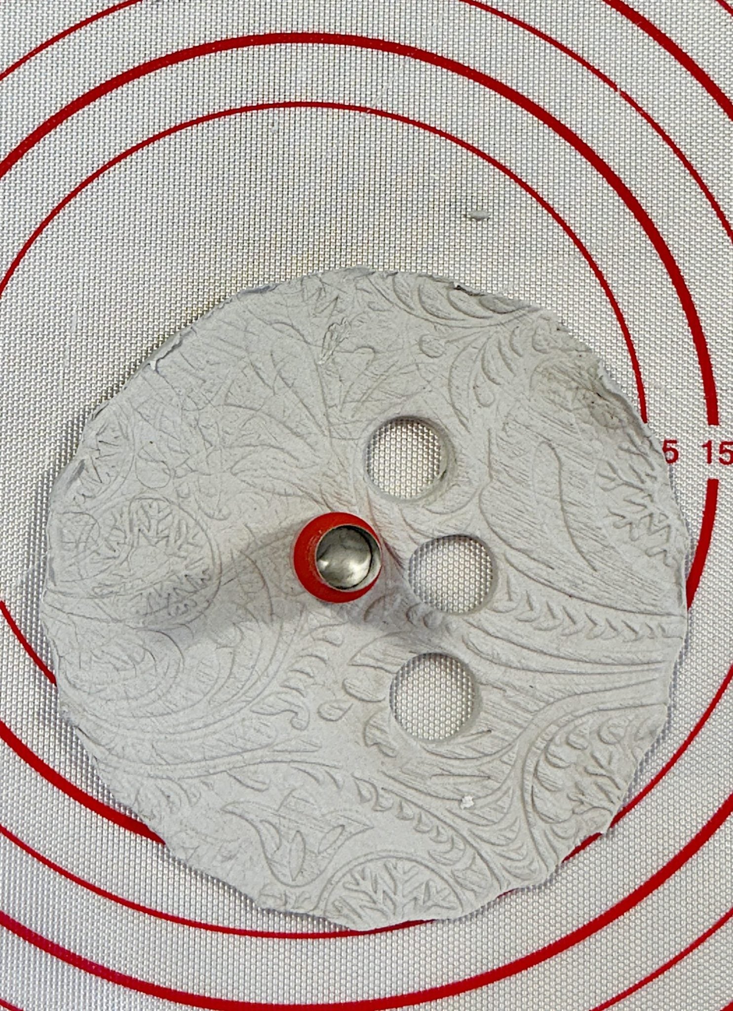 A textured clay circle on a silicone mat with concentric red circles, featuring three holes cut out using a red-handled tool.