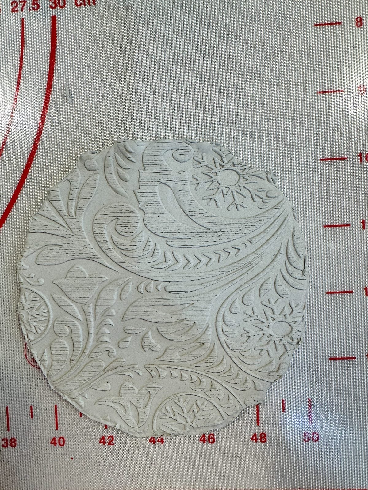Round piece of clay with intricate floral and paisley patterns on a textured surface featuring red measurement lines.