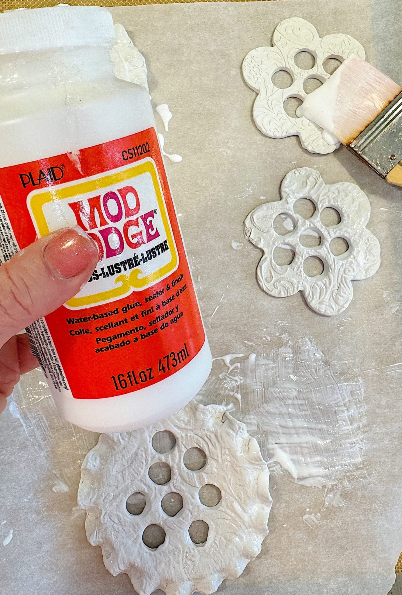 Hand holding Mod Podge bottle above clay or dough ornaments on a table, with a paintbrush nearby.