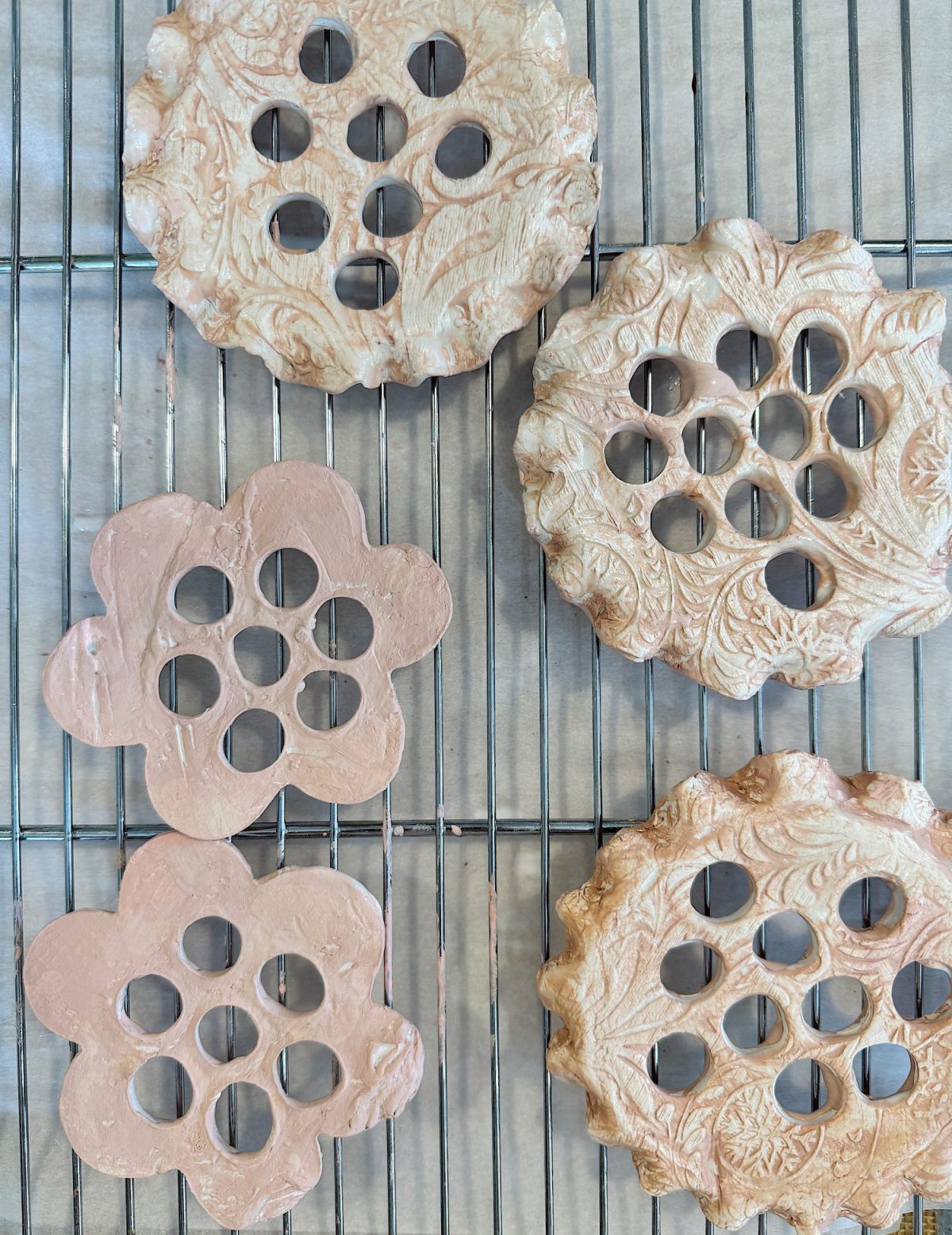 Ceramic flower-shaped pieces with embossed designs and multiple circular cutouts drying on a wire rack.