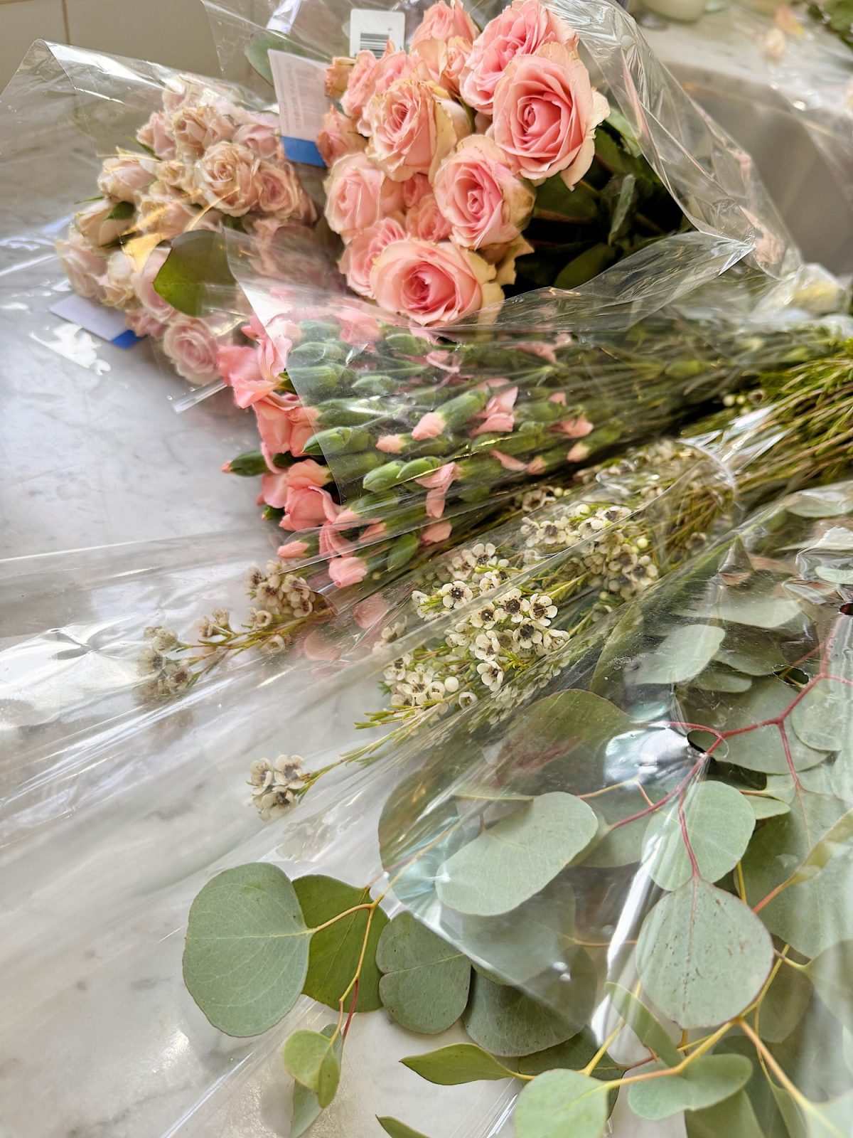 Bundles of pink roses, buds, and eucalyptus leaves wrapped in plastic lie on a marble surface.