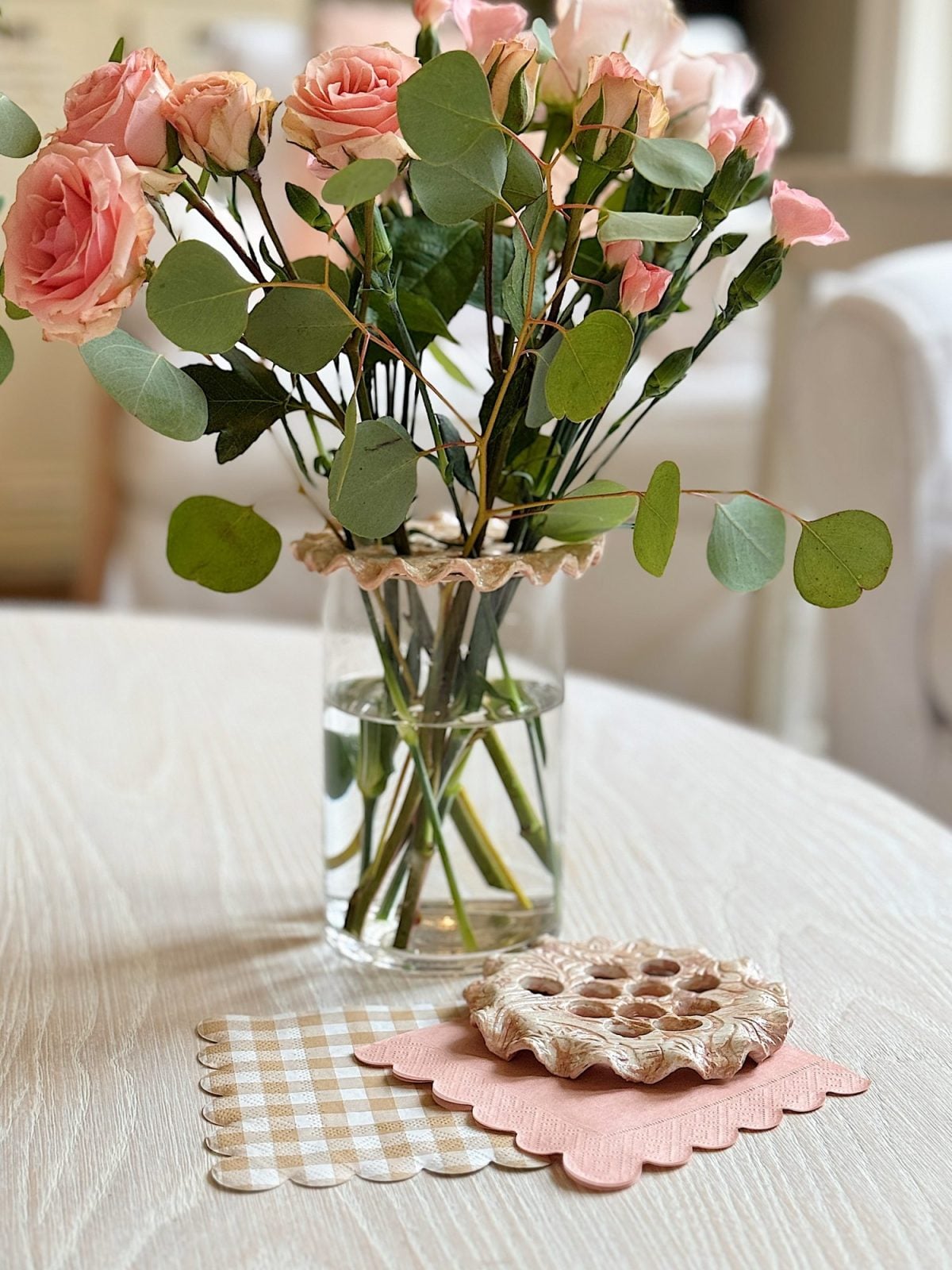 A bouquet of pink roses and eucalyptus in a glass vase on a table, alongside crocheted coasters and a gingham cloth.