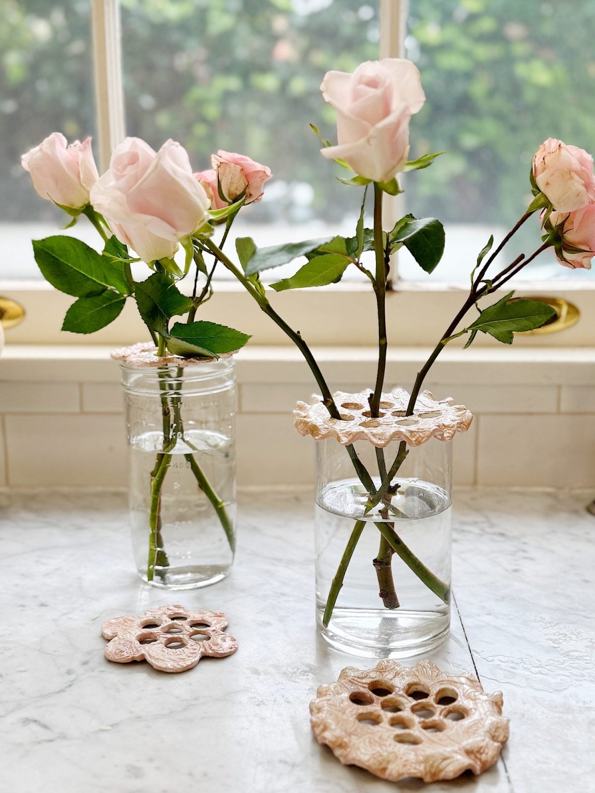 Two glass jars with pink roses placed in water on a marble surface. Each jar has a decorative floral-patterned holder. A similar holder is resting on the marble. Background shows a window.