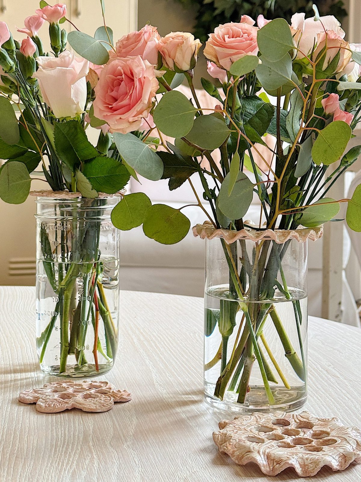 Two glass vases with pink and peach roses and green foliage on a light wooden table. Coasters with floral designs are placed under the vases.