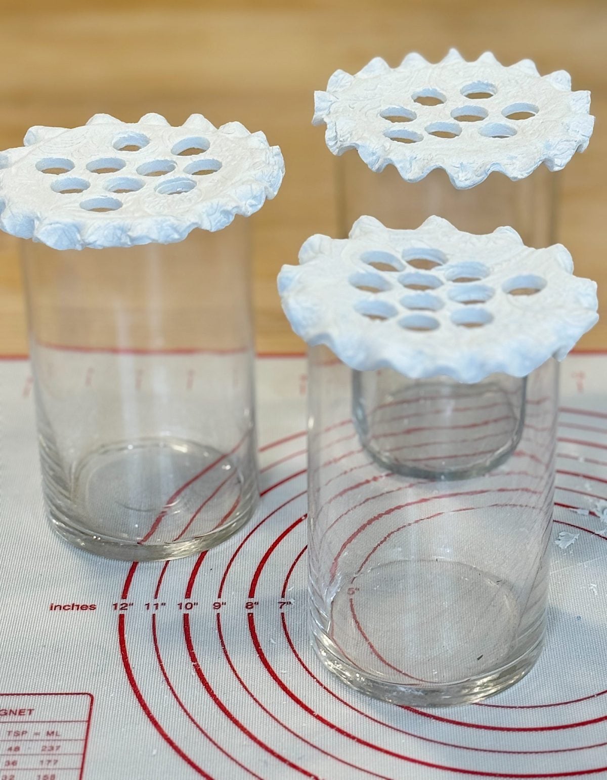 Three glass jars covered with white round fabric or paper filters featuring multiple holes, placed on a mat with red circular measurement markings.