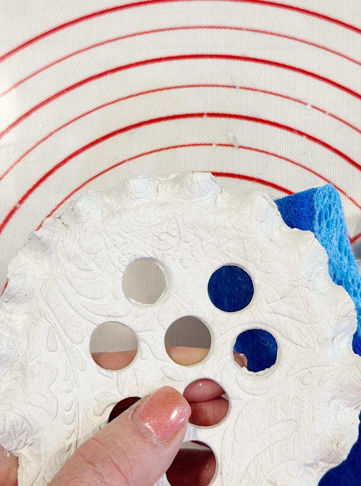 Hand holding a circular piece of clay with intricate designs and four holes, next to a blue sponge. Background has red concentric circles on a white surface.