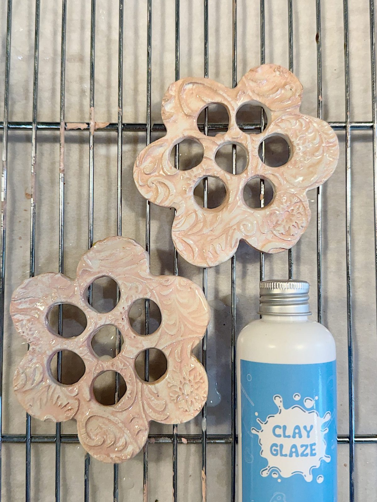 Two clay flower-shaped creations with circular cutouts are on a drying rack beside a blue bottle labeled "Clay Glaze.