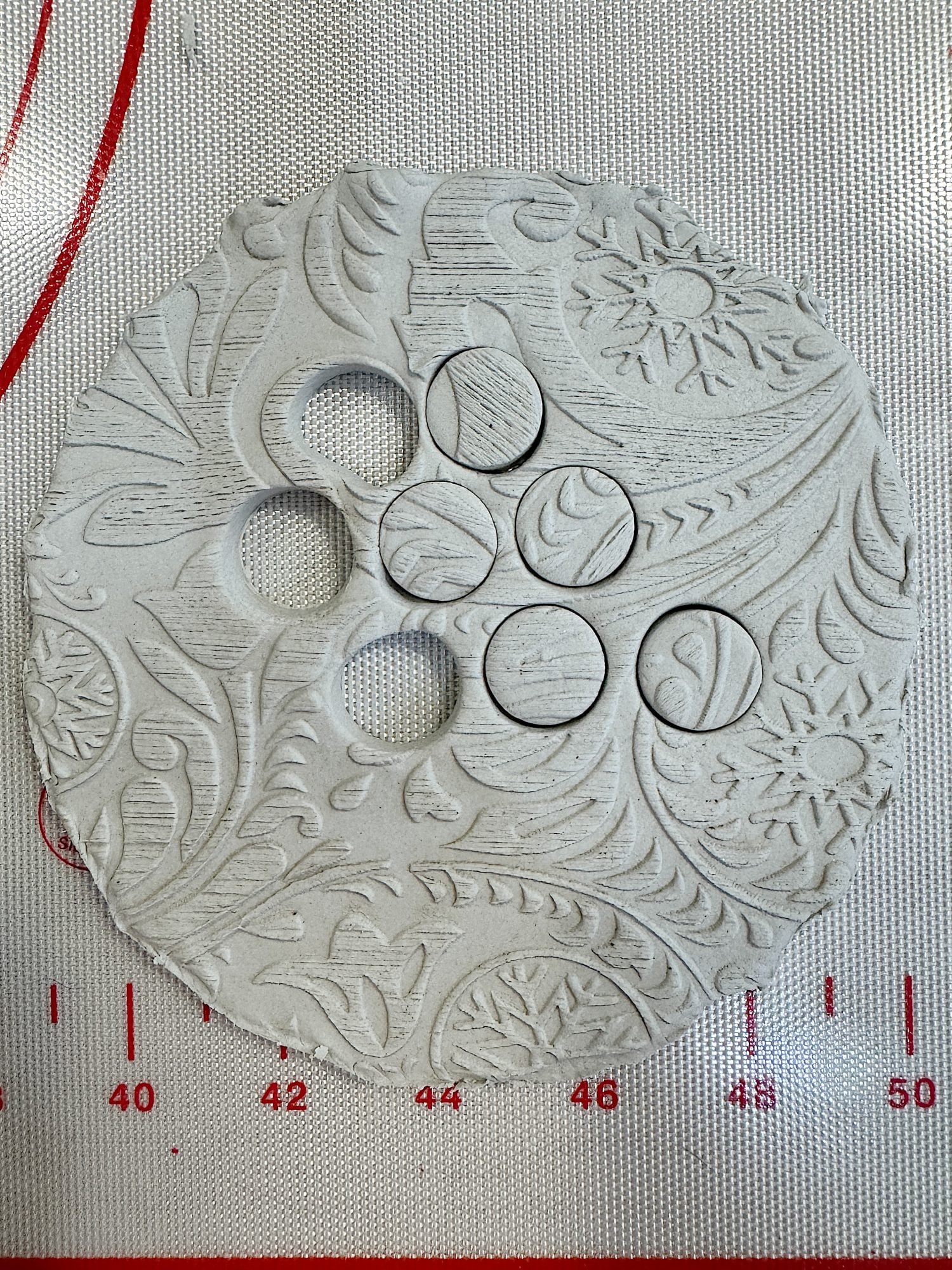 Textured clay circle with intricate floral and swirl designs, and six circular cut-outs of varying sizes on a gridded mat.