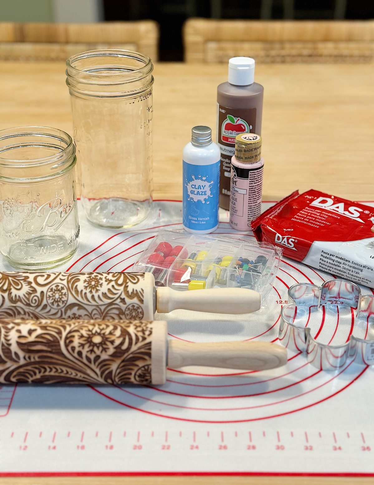 Crafting supplies on a table include mason jars, clay glaze, paints, clay, cookie cutters, and patterned rolling pins on a silicone mat.