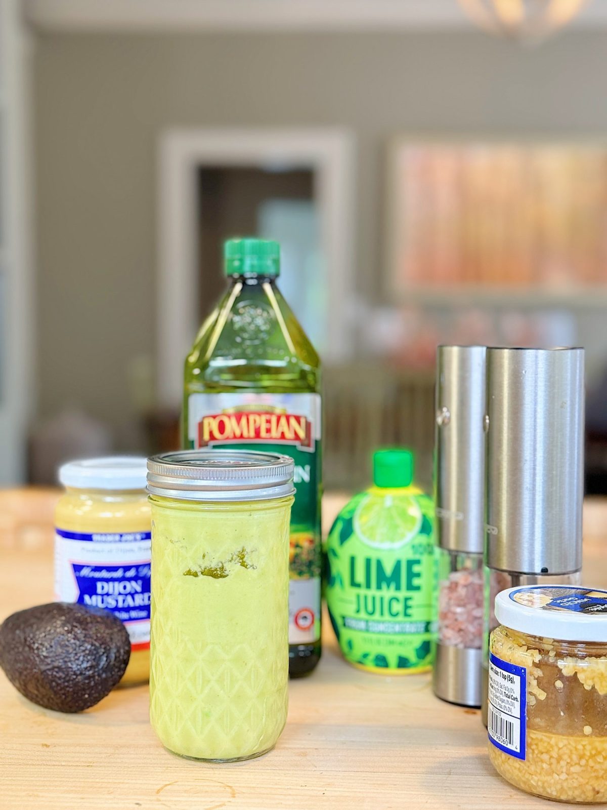 A jar of light green sauce, avocado, garlic, dijon mustard, olive oil, lime juice, salt, and pepper shakers on a wooden surface in a kitchen setting.