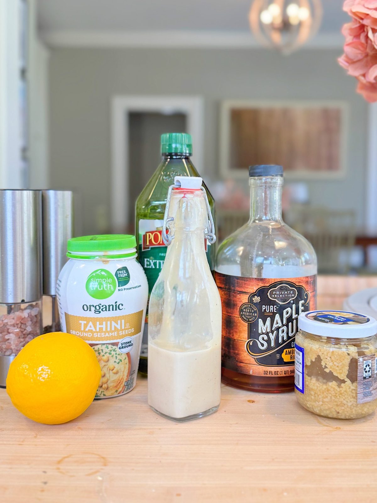 A group of kitchen items on a table, including tahini, olive oil, maple syrup, a lemon, salt, minced garlic, and a glass bottle with sauce.
