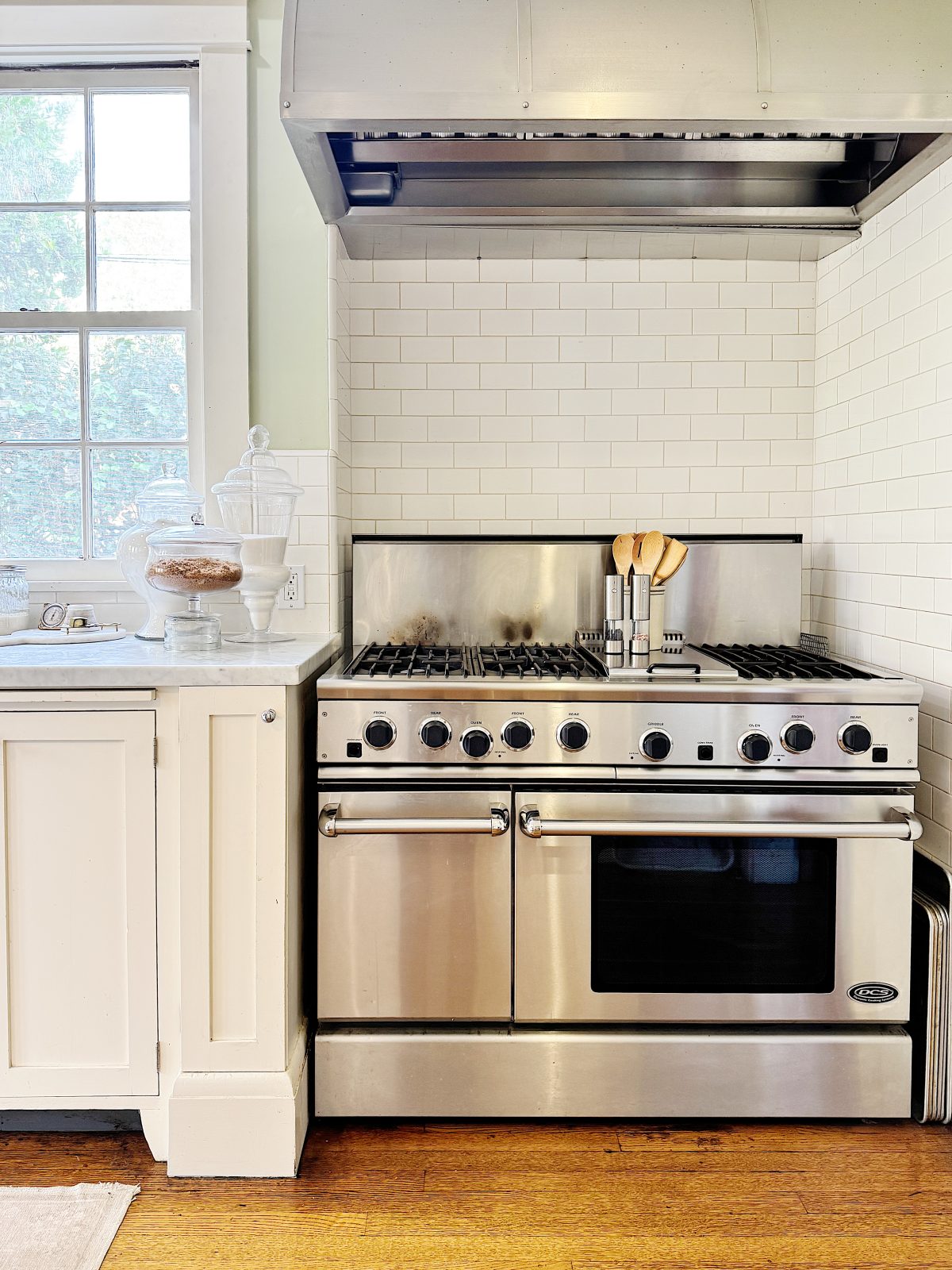 A stainless steel oven with a gas stovetop and hood, set against white subway tile. Kitchen utensils are in a holder on the stove, with jars on the counter.