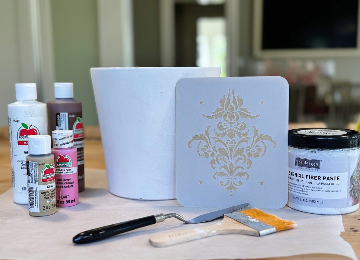 Assortment of art supplies on a table, including paint bottles, a white pot, a stencil with a floral design, a container of stencil fiber paste, a palette knife, and a paintbrush.