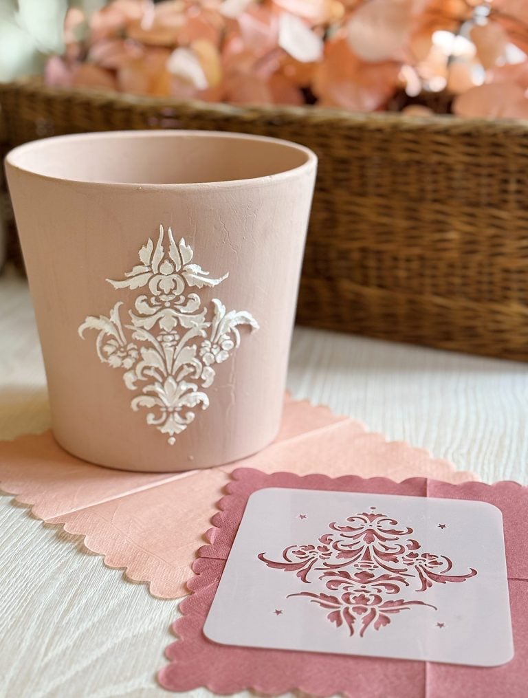 A pink plant pot with a white ornate design stencil. It is on a table with a matching pink napkin, and a wicker basket with dried leaves is in the background.