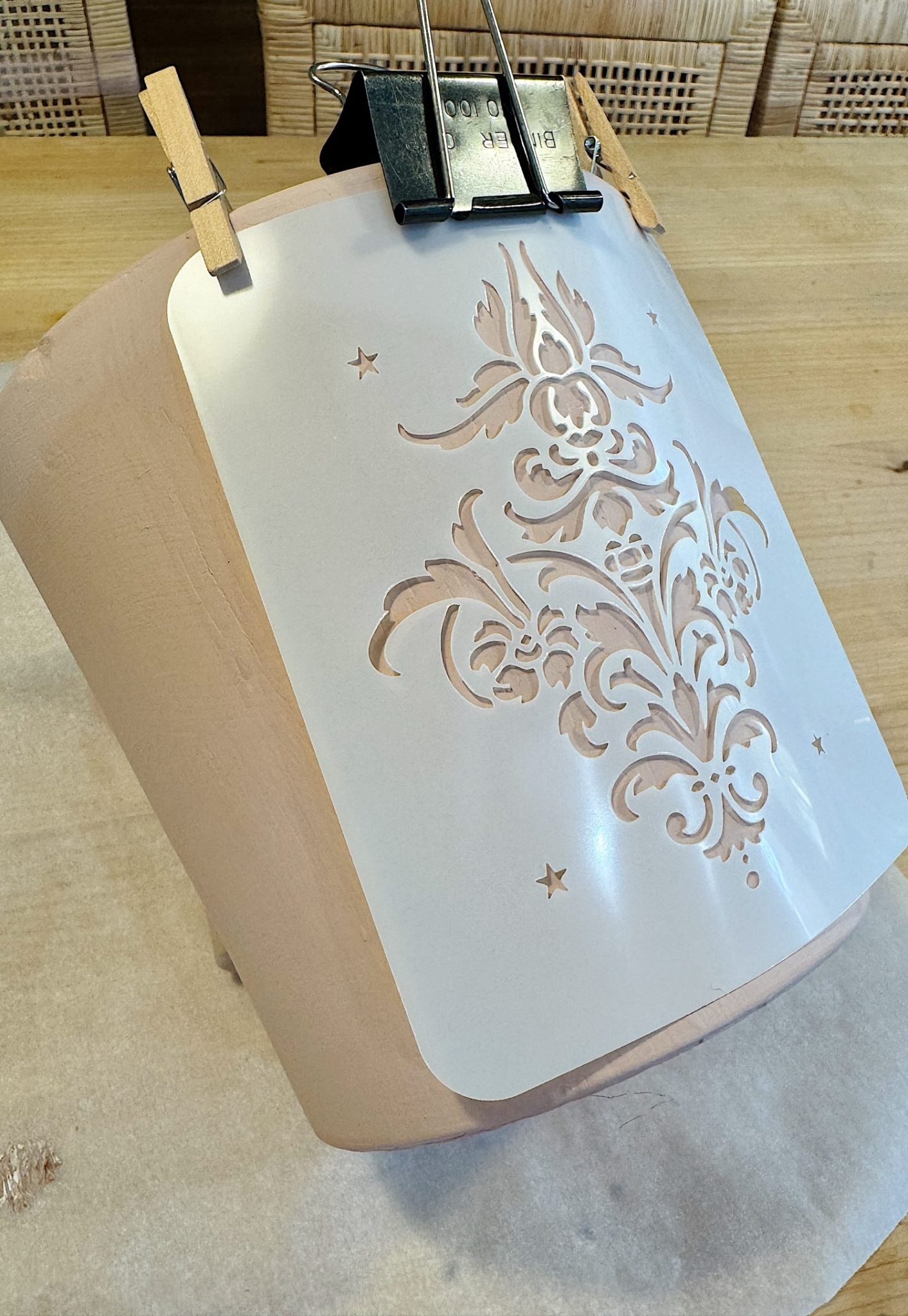 A wooden surface with an ornate stencil design held in place by two clothespins and a clip. The stencil features a symmetrical pattern with small stars.