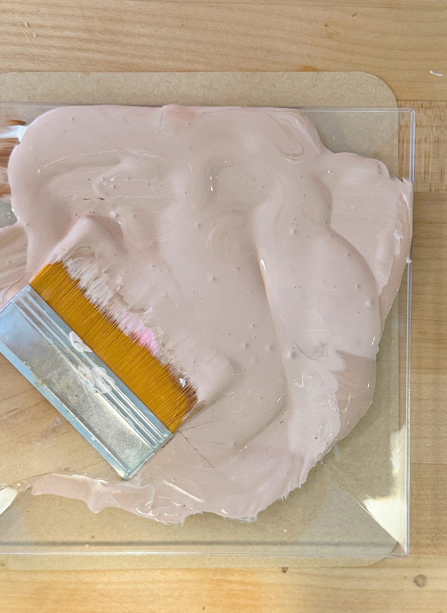 A paintbrush lies on a swirl of light pink paint spread across a clear plastic sheet on a wooden surface.