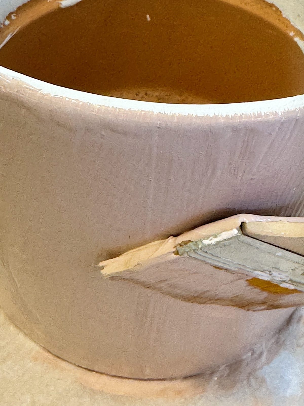 A mug being painted with a light brown paint using a brush.