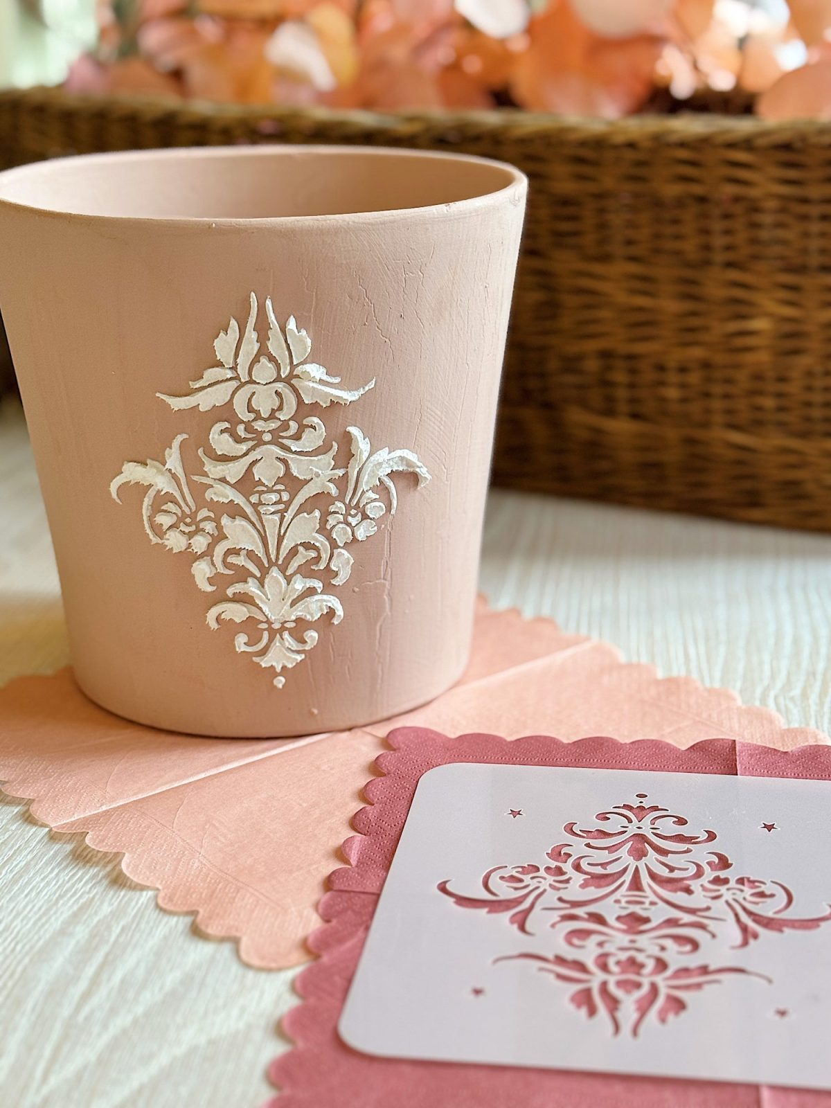 Beige pot with white floral pattern next to a pink napkin and card, both with matching designs, on a light wood surface.