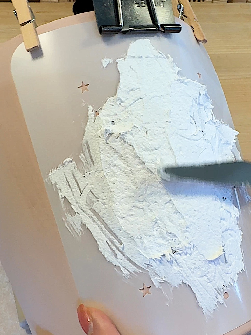 A hand is applying white plaster onto a plastic stencil with cut-out stars, using a spatula.