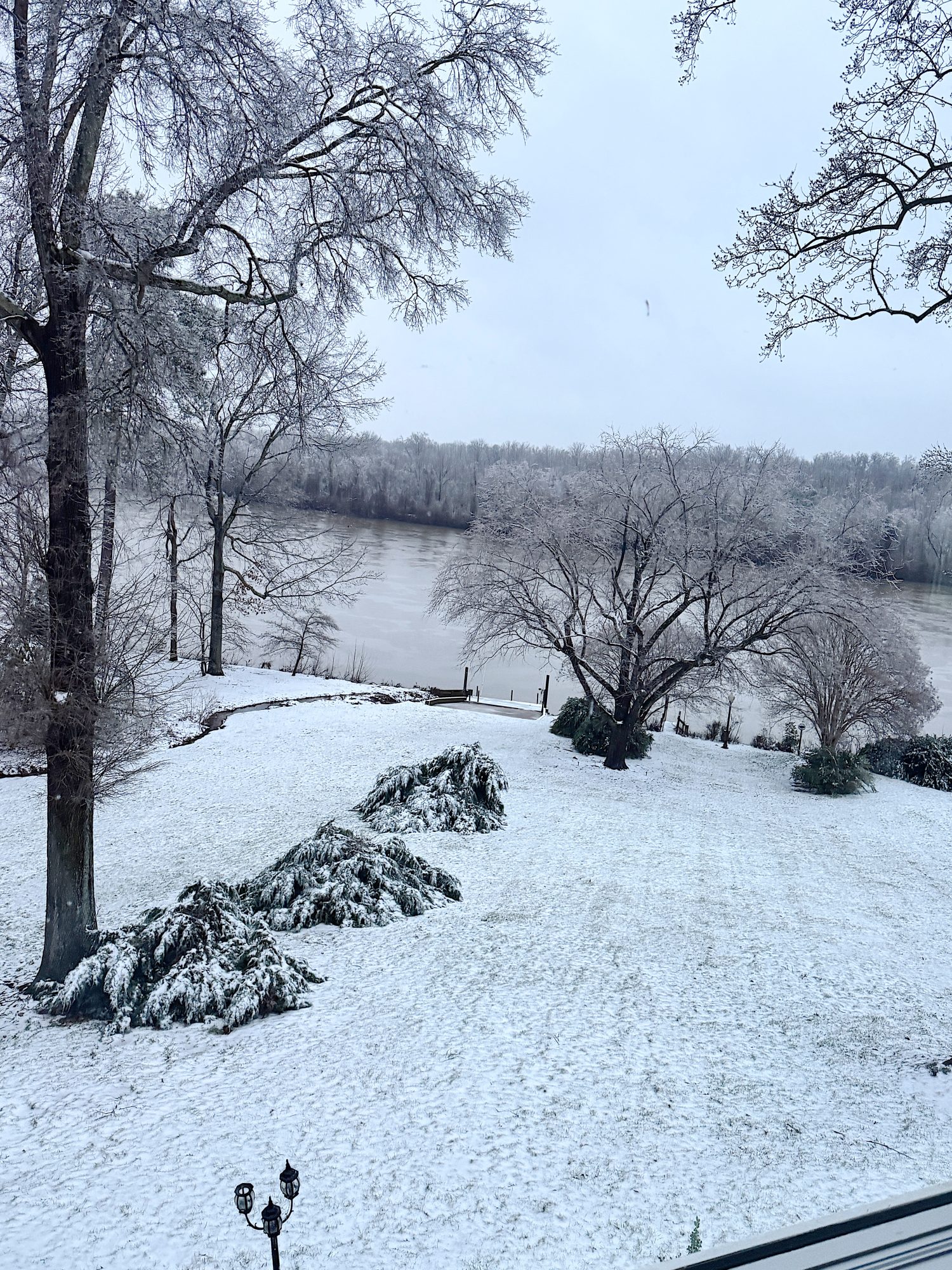 Snow-covered landscape with trees and a river in the background. Bare branches and frosty ground create a serene winter scene.
