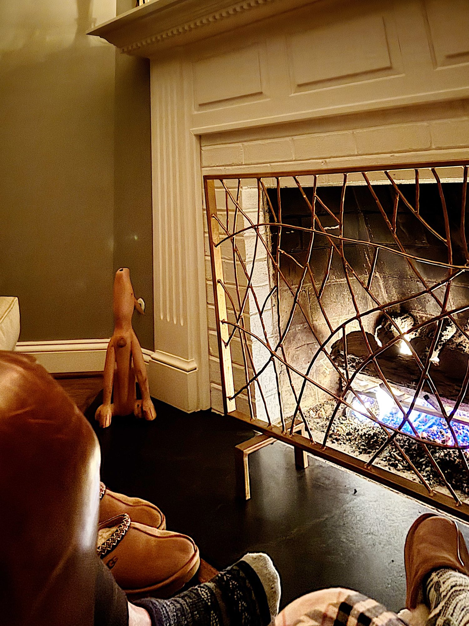Room with a lit fireplace, wooden dog sculpture on the floor, and a person in socks and plaid pants resting on a chair.
