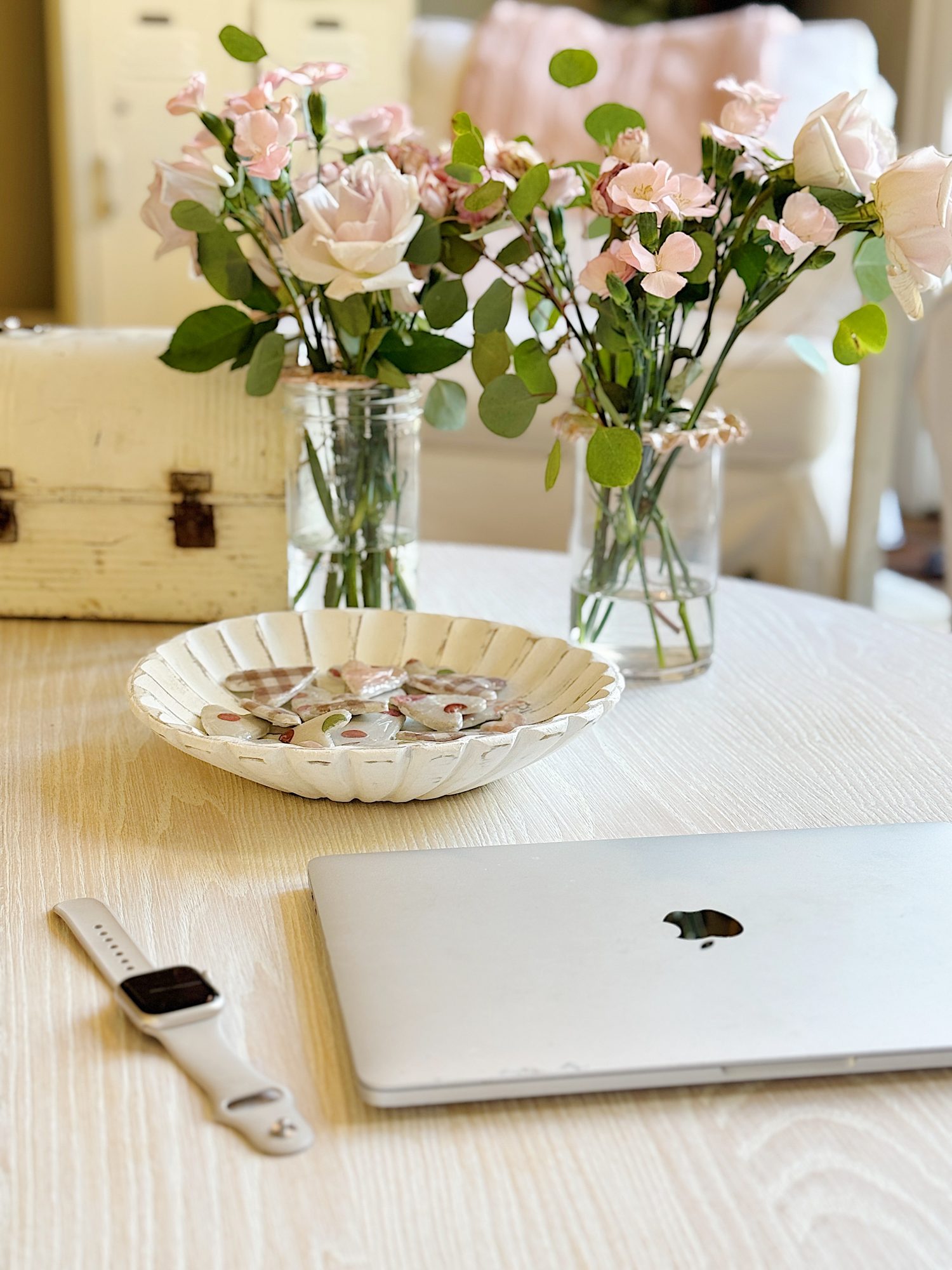 Laptop, smartwatch, and a decorative bowl on a light wooden table. Two jars with pink roses and greenery are in the background.