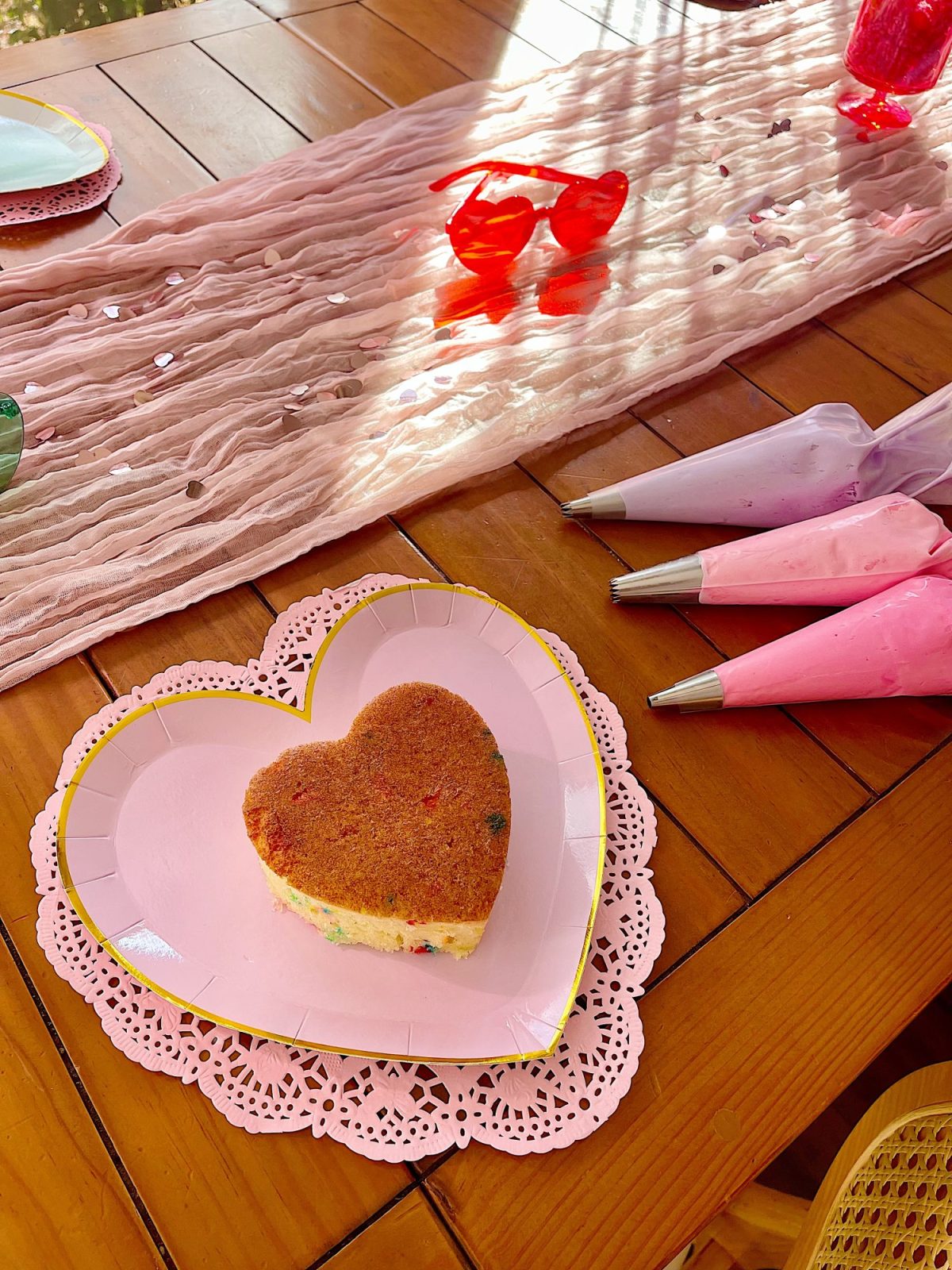 Heart-shaped cookie on a pink heart plate, surrounded by pink and purple frosting bags, with red glasses and a pink table runner in the background.