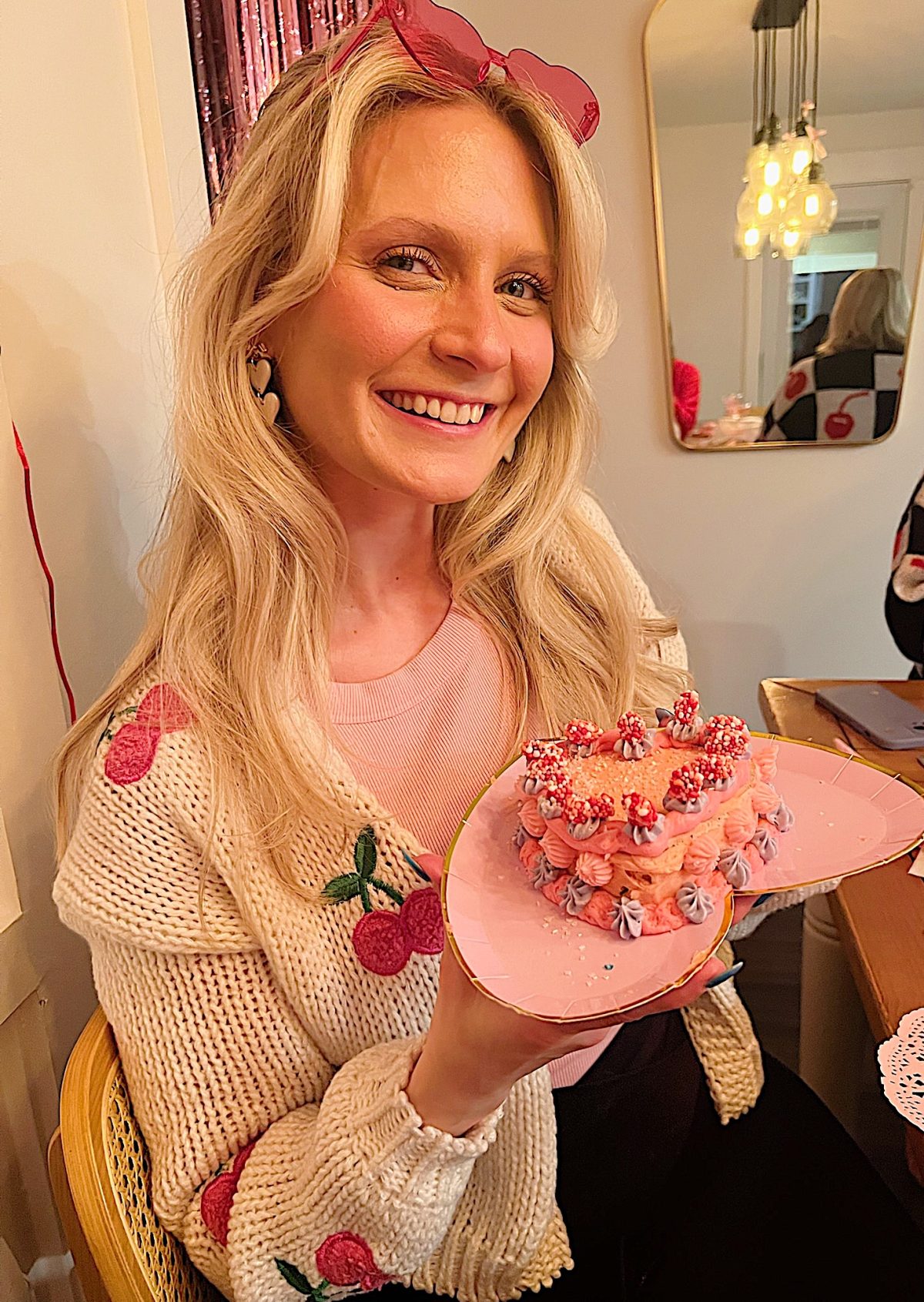 Woman smiling and holding a small pink cake with decorations, wearing a cherry-patterned sweater.