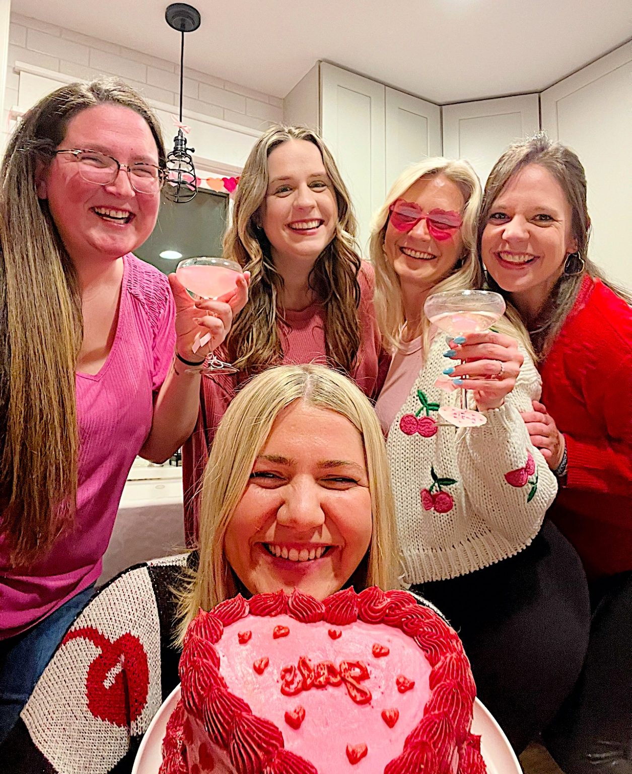 Five women smiling with drinks, one holding a heart-shaped pink cake with red icing. They are in a kitchen setting.