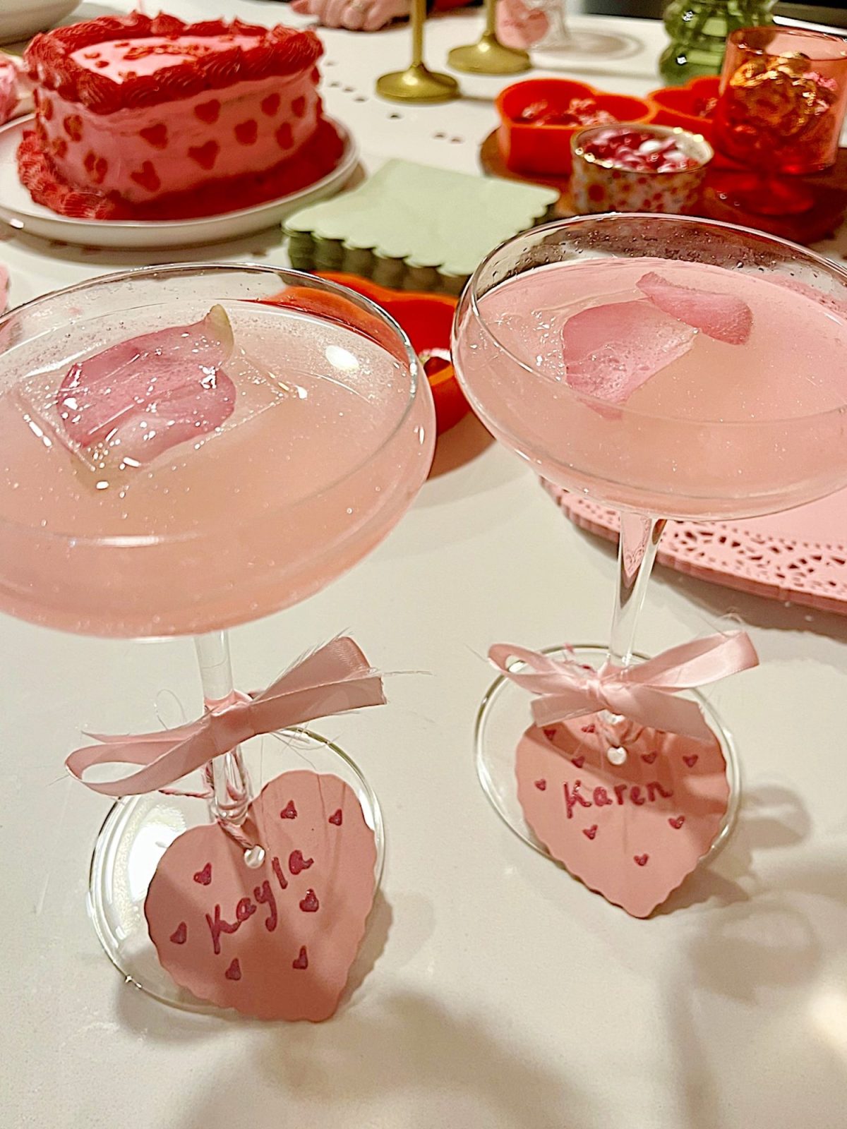 Two pink cocktails with rose petals, tagged "Kayla" and "Karen," on a table with a pink cake and various red and pink decorations.