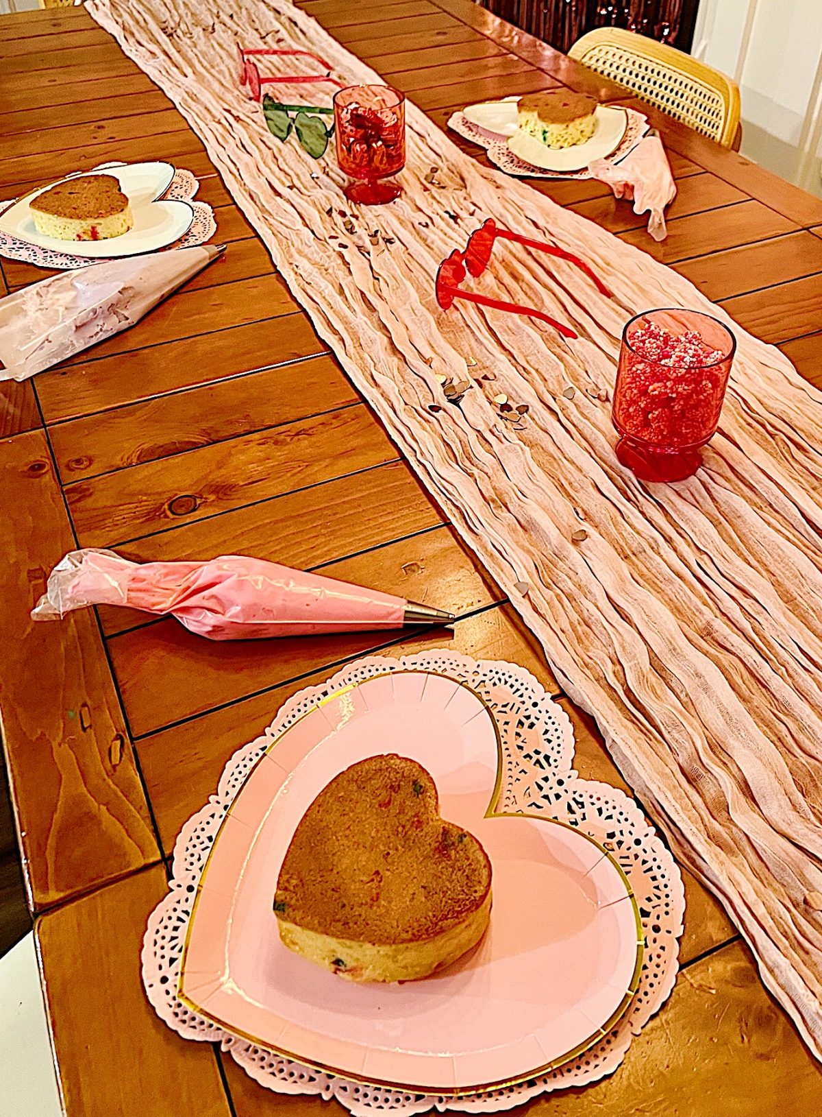 Wooden table set with heart-shaped plates and cookies, a pink fabric runner, and pink-tinted glasses filled with red snacks. Pastry bags with pink frosting are placed beside the plates.