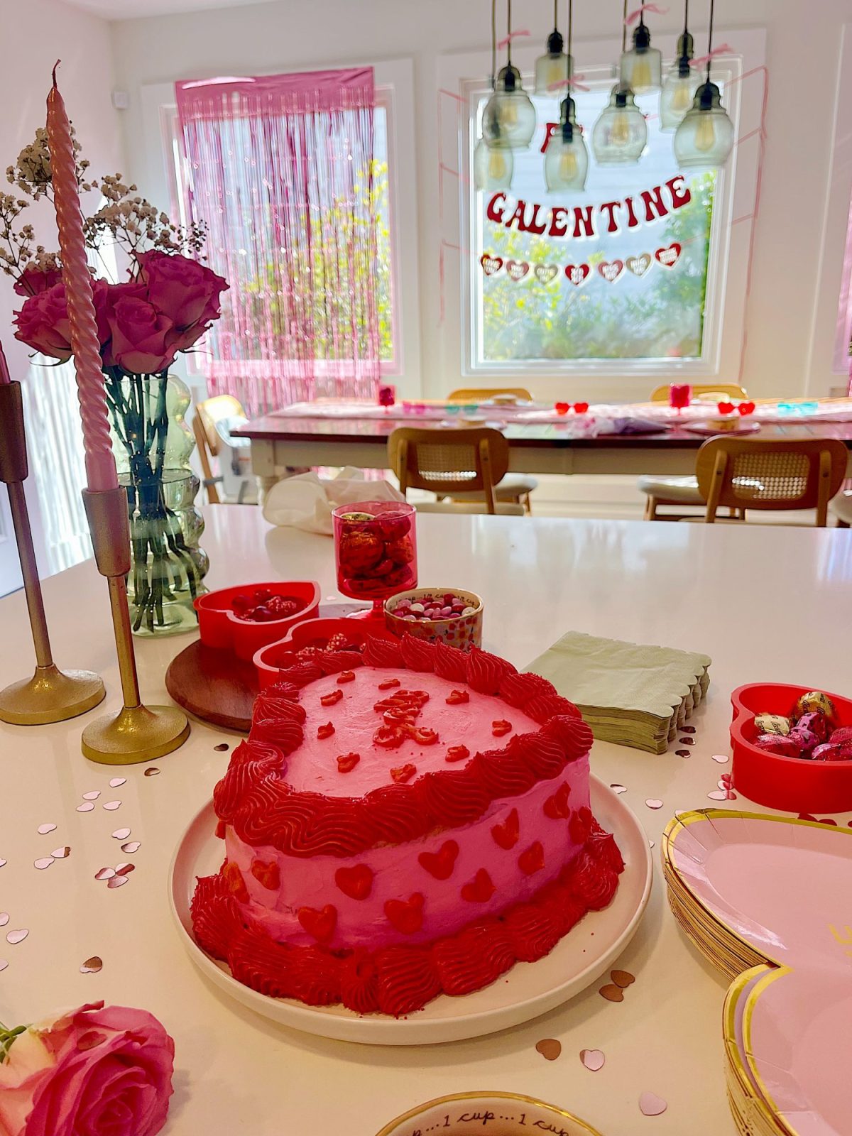 Pink and red heart-themed cake on a table with Galentine decorations, surrounded by candles, roses, and heart-shaped bowls.