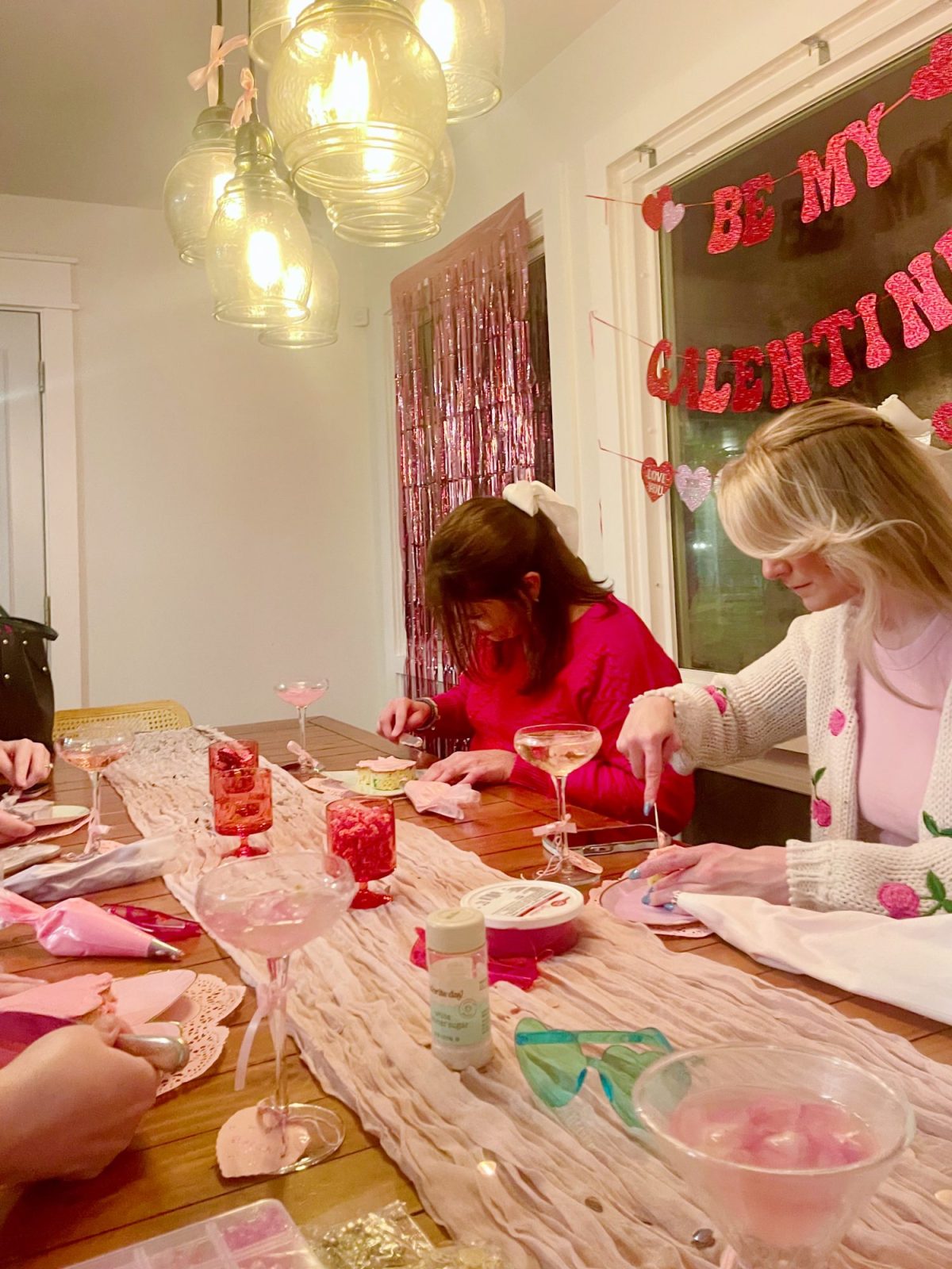 People sitting at a table decorated with Valentine's Day-themed items, crafting with heart-shaped materials. Hanging lights and a "Be My Valentine" banner are in the background.