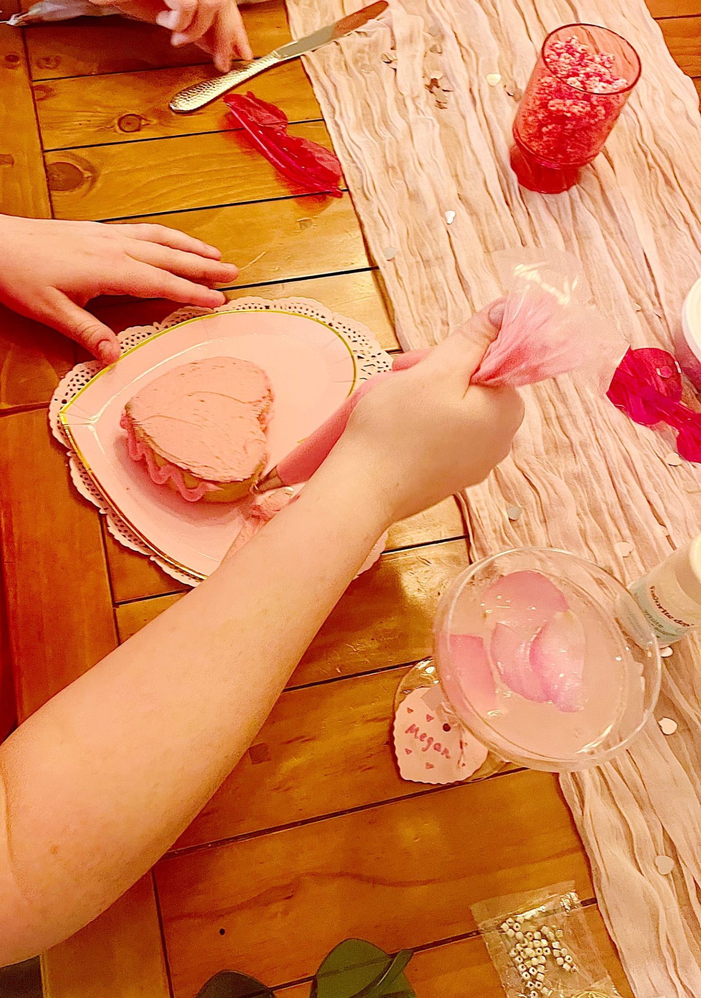 Person decorating a heart-shaped cake with pink frosting on a table set with drinks and decorations.
