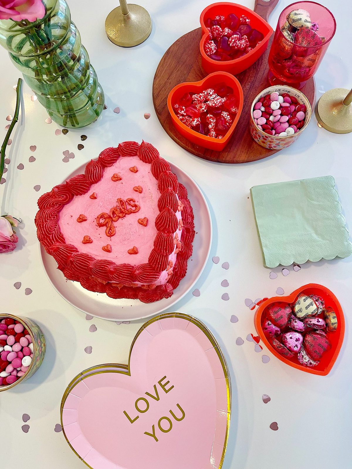 Heart-shaped cake with "Love" icing, surrounded by heart candies in red containers, a "Love You" plate, and scattered pink confetti on a white table.