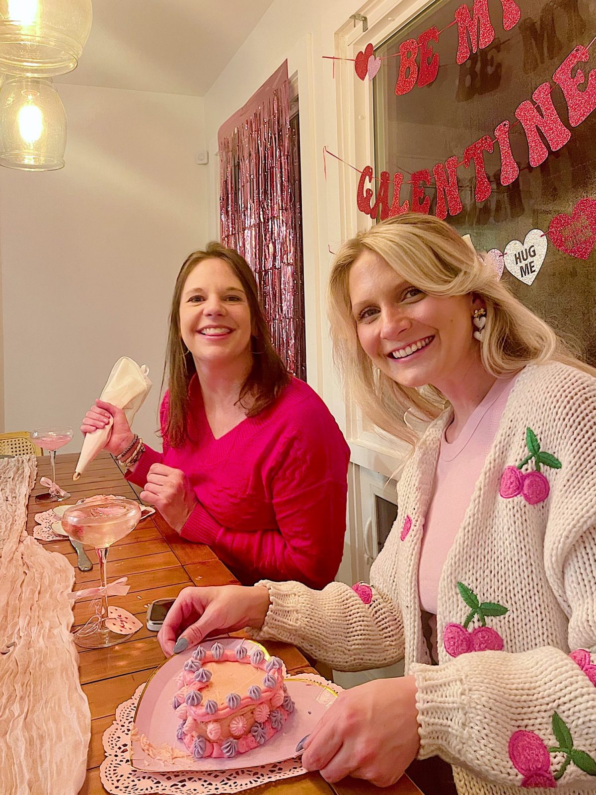 Two women sitting at a decorated table for Valentine's Day, one holding a piping bag and the other holding a spoon, with drinks and desserts in front of them.
